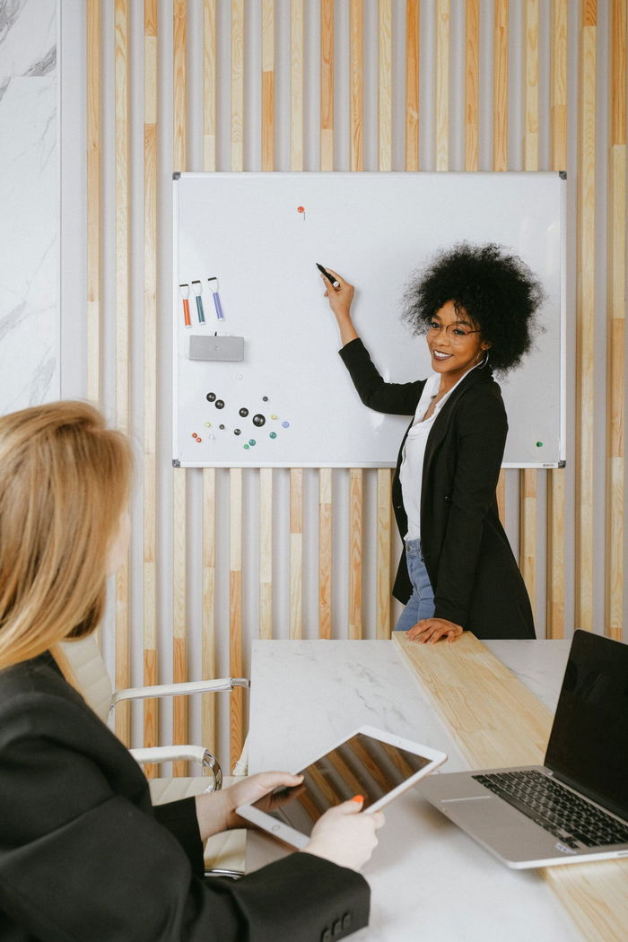 Woman writing on whiteboard in an office while another woman sits at a table holding a tablet.