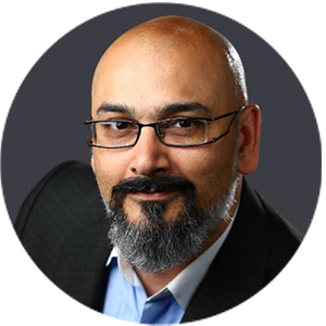 A person with glasses, a beard, and a bald head is wearing a dark suit jacket and light-colored shirt, set against a plain dark background.