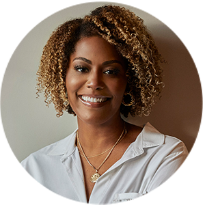 A person with curly hair smiles at the camera, wearing a white shirt and layered necklaces against a neutral background.
