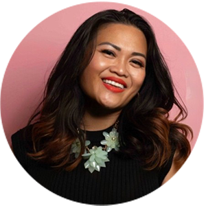 Person smiling, wearing a black top and a statement necklace, with a pink background.