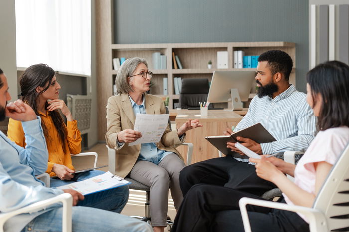 A group of people sitting in chairs in an office setting, engaged in discussion. One person is holding papers, possibly leading the meeting. Shelves with binders are in the background.