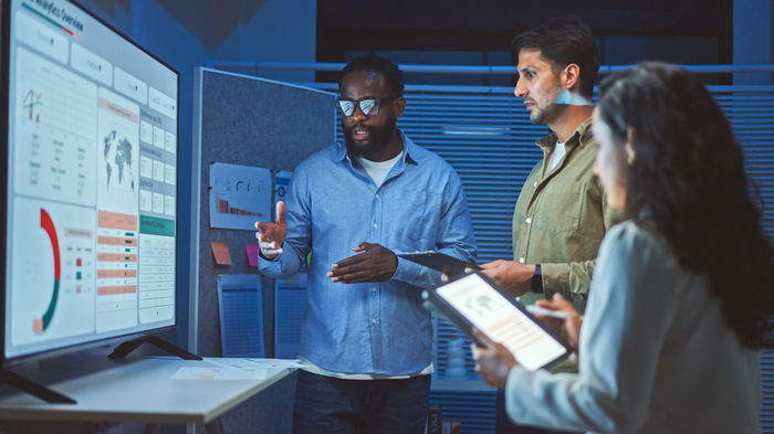Three people in an office setting review data on a large screen. The man in front gestures towards charts and graphs. Another man and a woman with a tablet listen attentively.