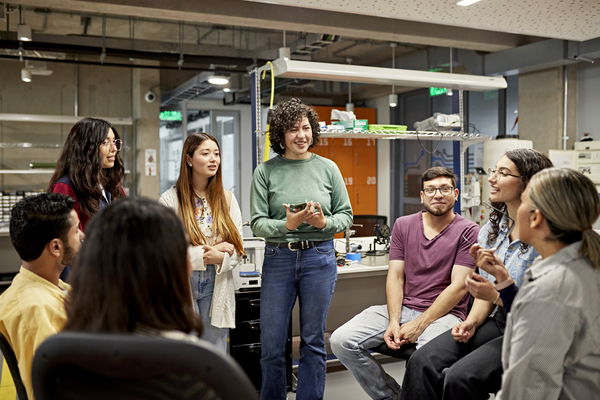 A group of people engaged in discussion, standing and sitting in a modern office or lab setting with various equipment on shelves in the background.