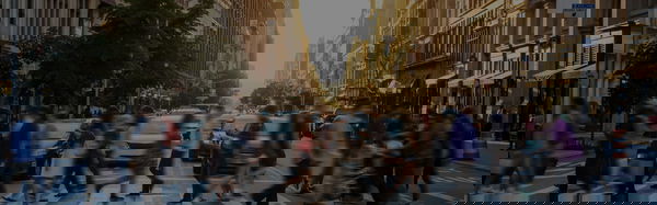 A busy city street with blurred pedestrians crossing at a crosswalk during sunset, surrounded by tall buildings and cars.
