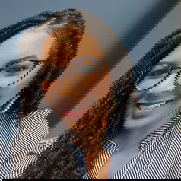 A woman with long curly hair, wearing glasses and a blue-and-white striped blouse, smiles confidently in front of a plain blue background—capturing the vibrant spirit of modern entrepreneurs.