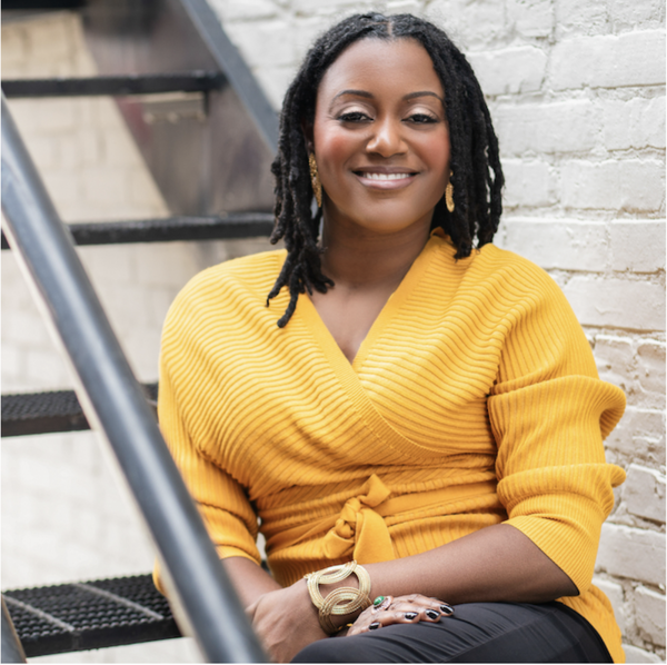 Bianca sits on a staircase next to a white brick wall, smiling at the camera. She is wearing a yellow wrap sweater and jewelry, and has medium-length dreadlocks.