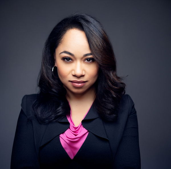 Jackie looks directly at the camera against a dark gray background. She has long dark hair, wearing a black blazer and a pink blouse.