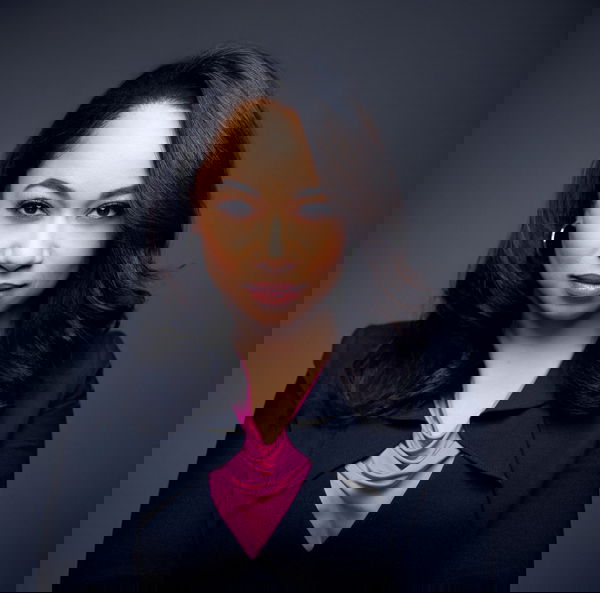 Jackie looks directly at the camera against a dark gray background. She has long dark hair, wearing a black blazer and a pink blouse.