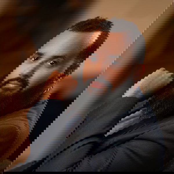 Carlos sits with his hand resting on his chin, looking at the camera. He has short brown hair, a beard and mustache. The background is softly blurred.