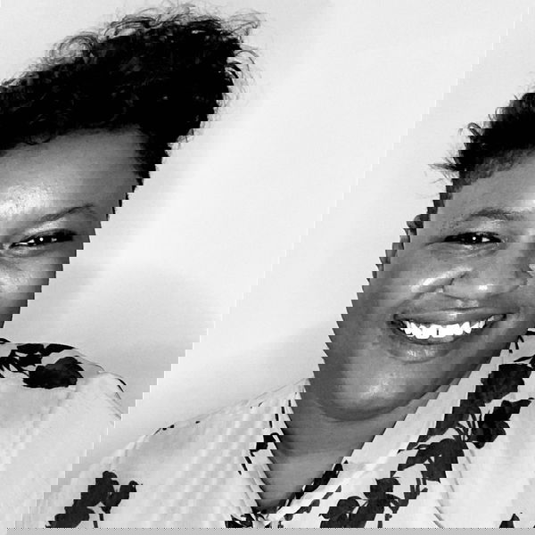 Joi has short curly hair and is smiling at the camera, wearing a light-colored top with a dark floral pattern. The photo is in black and white.