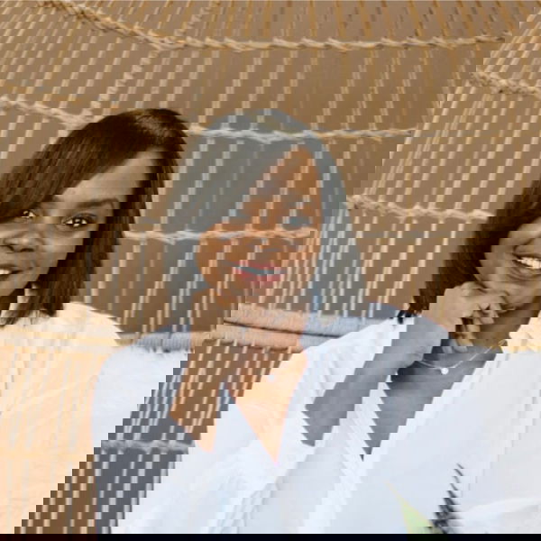 Katrina sits in front of a woven rope chair, smiling with her hand resting on her chin. She has shoulder-length dark hair and wears a white blouse.