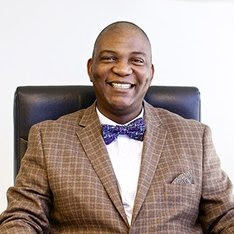 Ola sits in an office chair wearing a brown plaid suit and blue bow tie, smiling at the camera against a plain background.