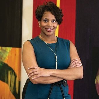 Penny stands with arms crossed in front of colorful abstract artwork. She has short hair and is wearing a teal sleeveless dress.