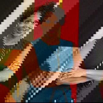 Penny stands with arms crossed in front of colorful abstract artwork. She has short hair and is wearing a teal sleeveless dress.