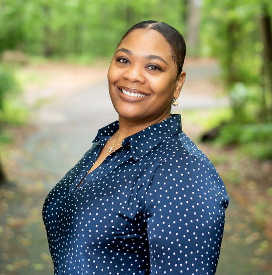 Raye is standing outdoors surrounded by greenery and smiling. She is wearing a navy blue polka dot blouse.
