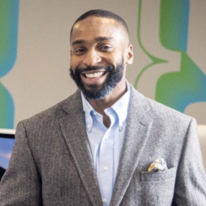 Waymond smiles while wearing a gray blazer, light blue shirt, and pocket square, standing indoors with a modern abstract background.