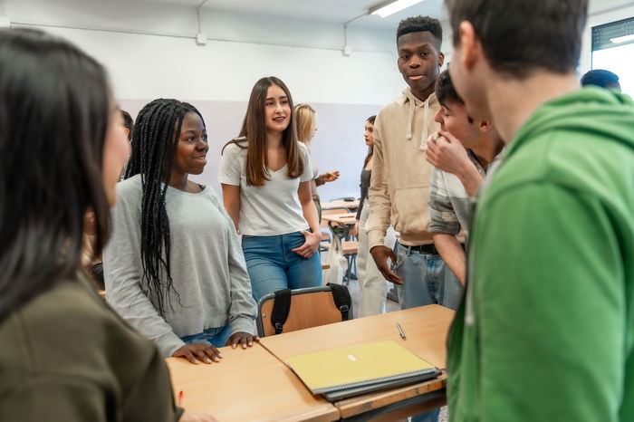 A group of students stand and talk around a desk in a classroom, with notebooks and folders on the table.