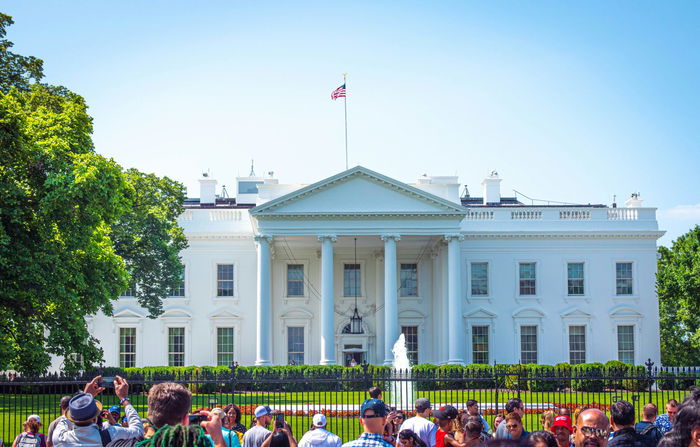 The White House, a white neoclassical building with an American flag on top, viewed from outside the front fence with a crowd of people in the foreground.
