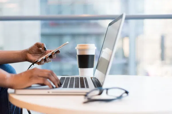 A person types on a laptop with one hand while holding a smartphone in the other; a coffee cup and eyeglasses rest on the table nearby.