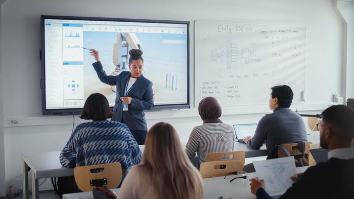 An instructor in business attire presents data on a large screen to a group of students seated in a classroom with laptops, while diagrams are visible on a whiteboard.