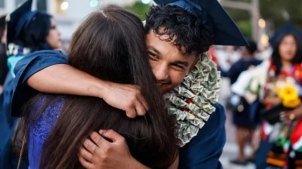 A graduate in a blue cap and gown, wearing a necklace made of dollar bills, hugs another person during a graduation ceremony.