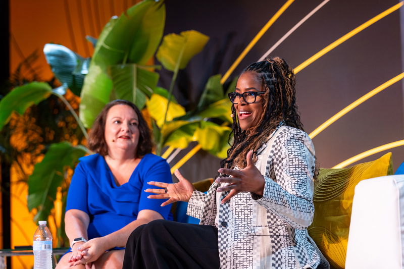 Two women sit on stage in conversation; one in a blue dress listens while the other, wearing a patterned jacket, speaks animatedly. Plants and yellow lines decorate the background.