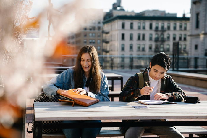 Two students sit at an outdoor table; one smiles while opening a bag, the other writes in a notebook. City buildings are visible in the background.