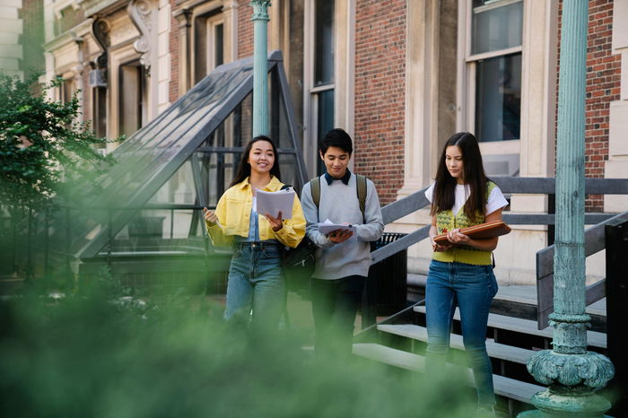 Three students walk down stairs outside a brick building, carrying books and backpacks, appearing to converse and read as they go.