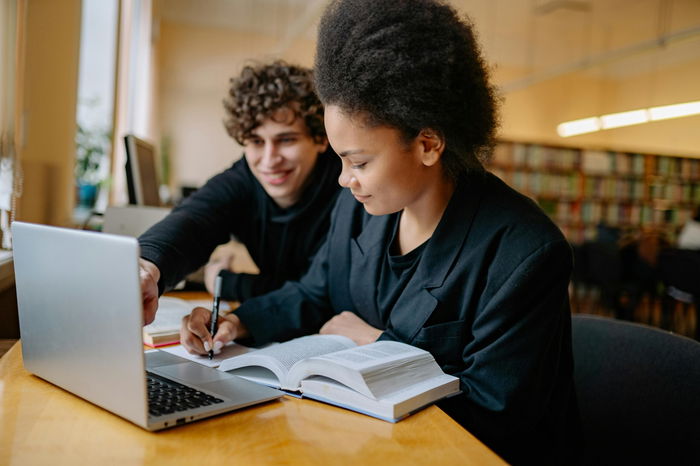 Two students study together at a table in a library, using a laptop and an open textbook. One points at the laptop screen while the other takes notes.