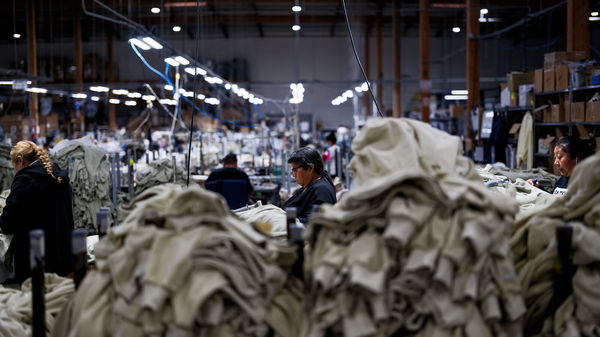Workers in a large factory sort and sew fabric surrounded by piles of beige-colored cloth under bright overhead lighting.