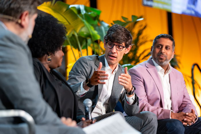 Four people sit on a panel discussion. One man in a gray suit gestures while speaking, with two others listening and one person holding papers. Green plants and orange lighting are in the background.