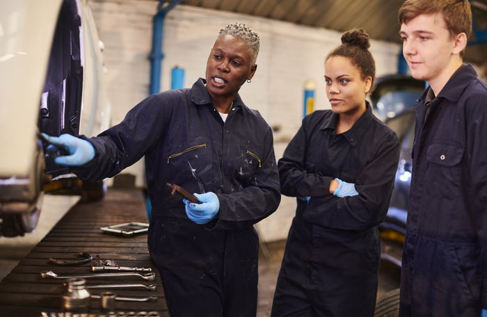 An instructor in coveralls explains car maintenance to two students in a workshop, pointing at parts under a raised vehicle. Tools are laid out on a bench nearby.