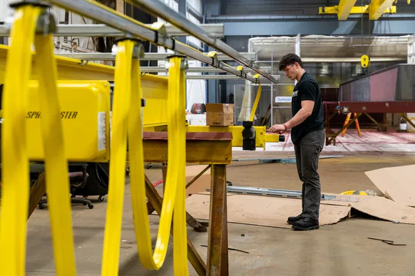 A man operates machinery using a control panel in an industrial workshop with yellow equipment and metal structures.