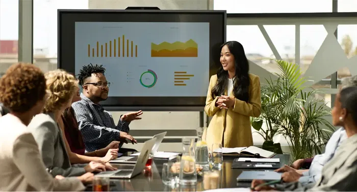 Six people sit around a conference table while a woman stands and presents charts and graphs displayed on a large screen in a modern office setting.