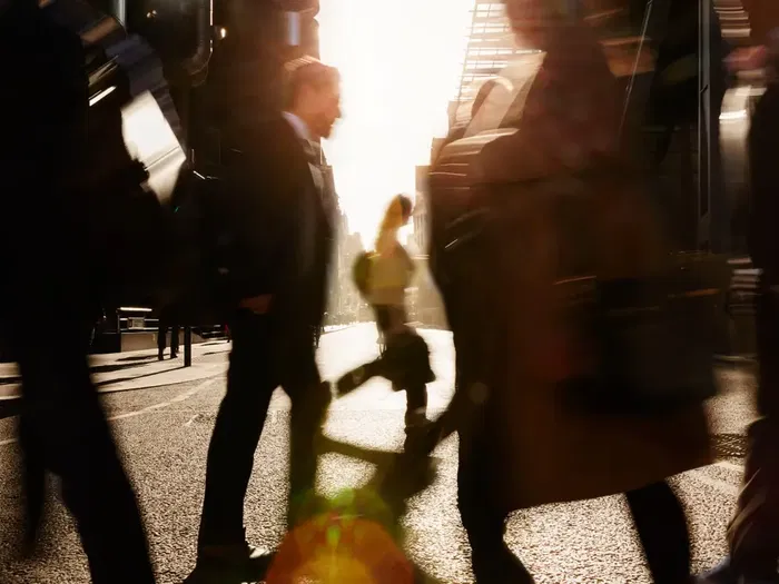 People walk across a city street in blurred motion, with sunlight streaming between tall buildings in the background.